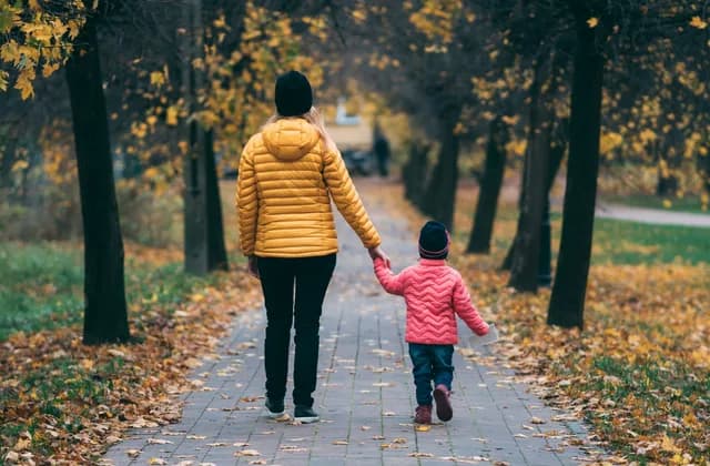 Adulto e criança de mãos dadas caminhando por uma alameda com folhas de outono, vistos de costas