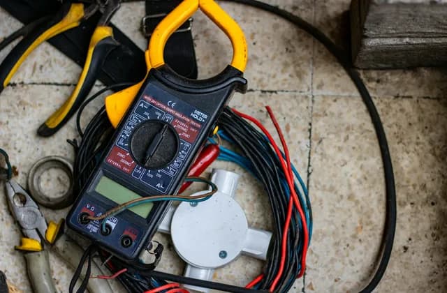 Alicate amperímetro, alicates, fios coloridos e uma caixa de passagem sobre um piso de ladrilho