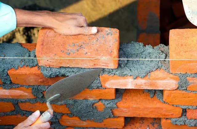 Mãos assentando tijolos cerâmicos com argamassa e colher de pedreiro, guiadas pela linha