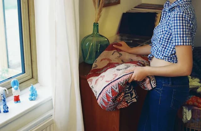 Mujer doblando una colcha estampada junto a un mueble de madera, en casa