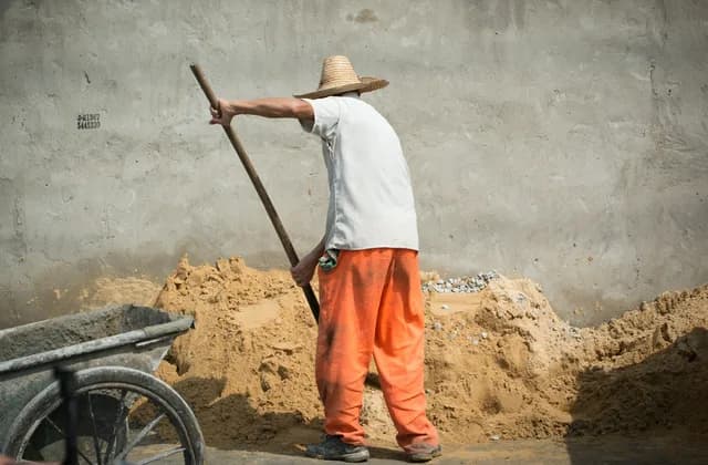 Albañil con sombrero de paja paleando arena junto a una carretilla en una obra
