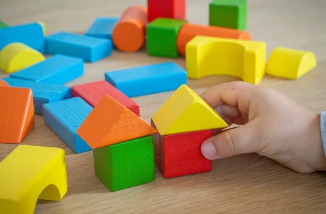 Mano de un niño colocando un bloque amarillo sobre una casita de bloques de madera de colores