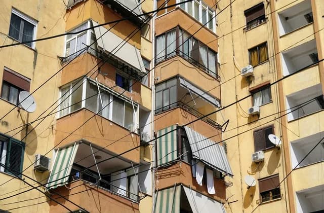 Güneş alan sarı cepheli bir apartmanda balkonlara asılı klima dış üniteleri, tenteler ve cephe boyunca uzanan kablolar