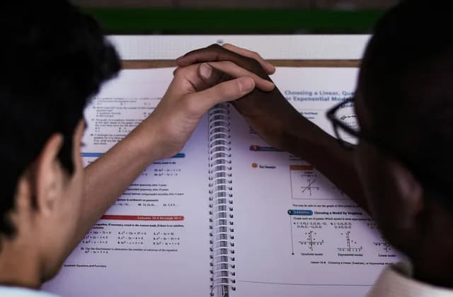 Bir öğrenci ve öğretmen açık ders kitabı üzerinde birlikte çalışıyor, yalnız eller ve defter görünüyor