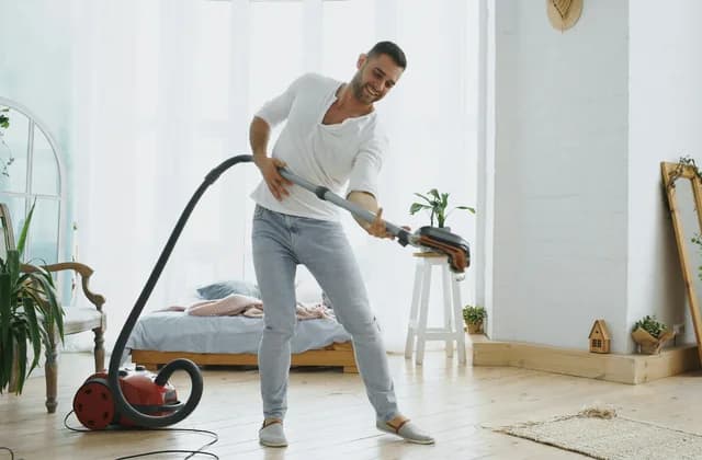 Man vacuuming the floor of a bright bedroom with a canister vacuum cleaner