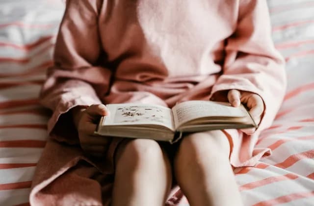 Child in a pink dress sitting on a bed holding an open storybook