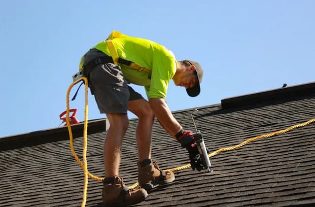 Roofer in a safety harness applying sealant on an asphalt shingle roof