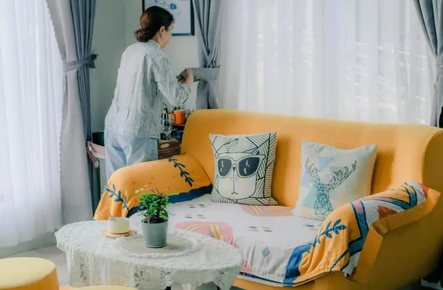 Mulher arrumando a cortina ao lado de um sofá amarelo coberto com manta e almofadas estampadas