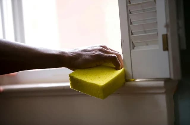 Mano con una esponja amarilla limpiando el marco de una ventana