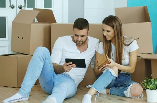 Pareja sentada en el piso entre cajas de mudanza revisando una tableta