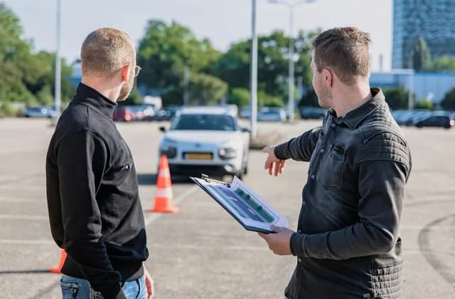 A driving instructor with a clipboard explaining a manoeuvre to a learner beside a traffic cone