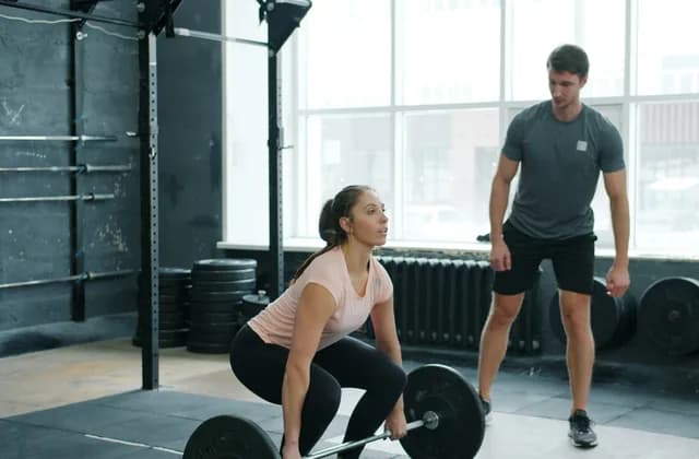 A personal trainer watching a client set up a barbell lift in a gym
