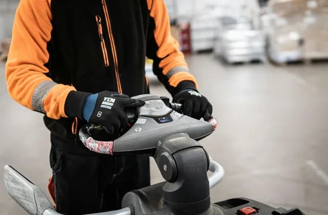 Gloved hands on the tiller controls of a powered pallet truck in a warehouse aisle