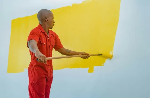 Pintor con overol rojo pintando de amarillo una pared blanca con un rodillo de mango largo