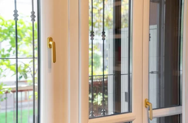 Ventana de PVC blanca con manilla dorada y reja de hierro al otro lado del cristal