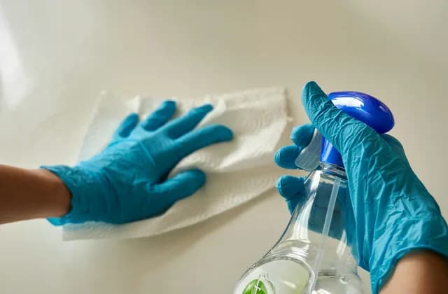 Manos con guantes azules limpiando una superficie blanca con un paño y un pulverizador
