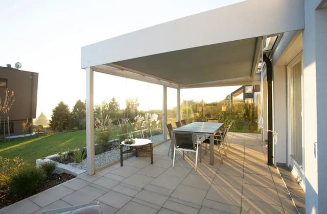 A covered patio with glass sides, a dining table and chairs, next to a sunny garden lawn with trees in late afternoon light