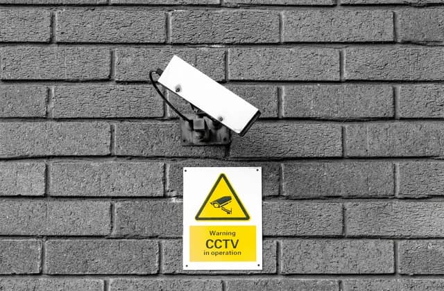 CCTV camera on a grey brick wall above a yellow warning sign