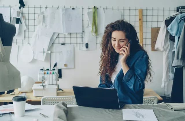 Dona de um ateliê de costura falando ao celular diante do notebook