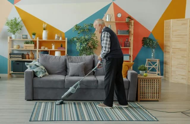 Ältere Person saugt einen gestreiften Teppich vor einem grauen Sofa im Wohnzimmer