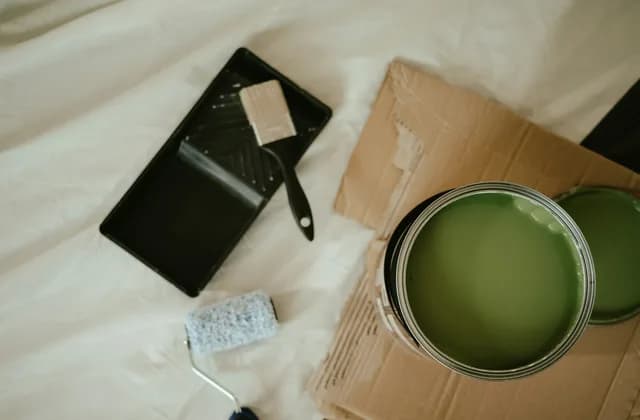 Pot de peinture verte ouvert, bac à peinture, pinceau et rouleau posés sur une bâche de protection