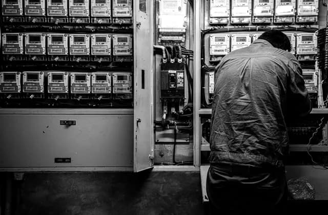 Sayaç ve pano dolaplarıyla dolu bir elektrik odasında, açık bir panonun önünde sırtı dönük çalışan elektrikçi; siyah beyaz kare