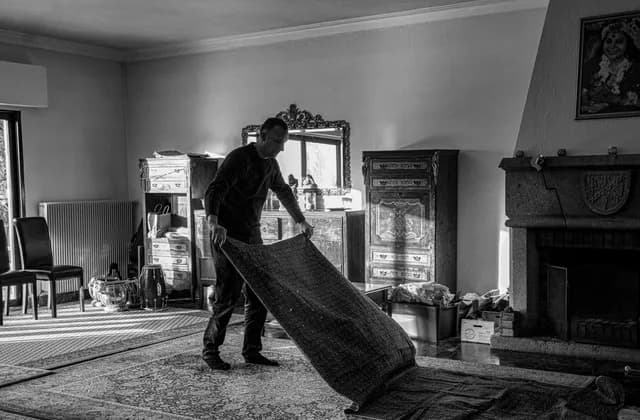 Mann rollt in einem ausgeräumten Wohnzimmer einen Teppich zusammen, Schwarzweißfoto