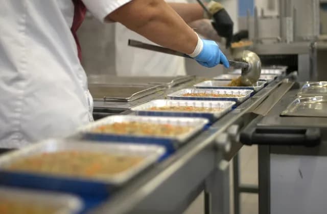 Person im weißen Kittel portioniert mit Handschuhen Essen in Menüschalen auf dem Band einer Großküche