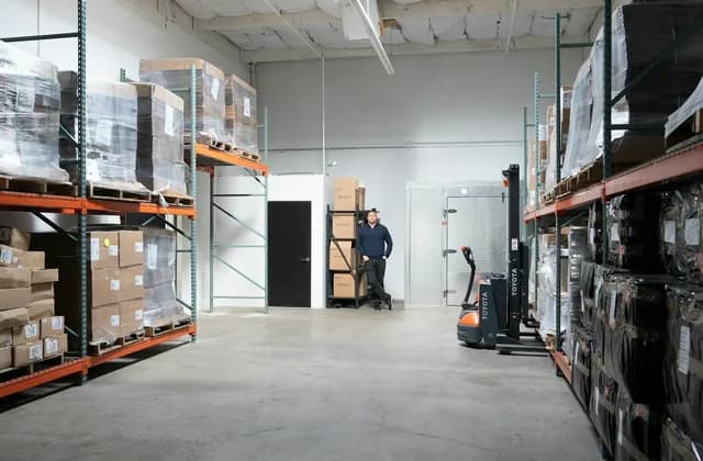 Lagerhalle mit Palettenregalen, im Hintergrund eine Person neben einem Elektro-Hubwagen