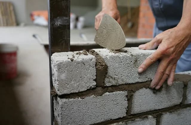A bricklayer’s hands spreading mortar with a trowel on a wall of grey cement blocks, next to a steel upright