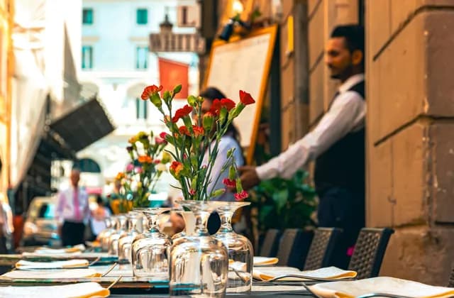 Gedeckter Tisch im Schanigarten mit Weingläsern und Nelken, dahinter ein Kellner mit Weste und weißem Hemd an der Speisekarte