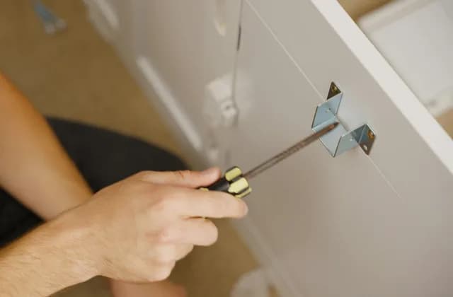 Monteur schraubt mit einem Schraubendreher einen Metallwinkel an eine weiße Schrankfront