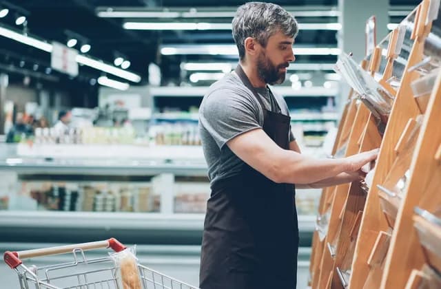 A shop worker in a dark apron checking a wooden bread display in a supermarket, with a baguette in a trolley in front of him