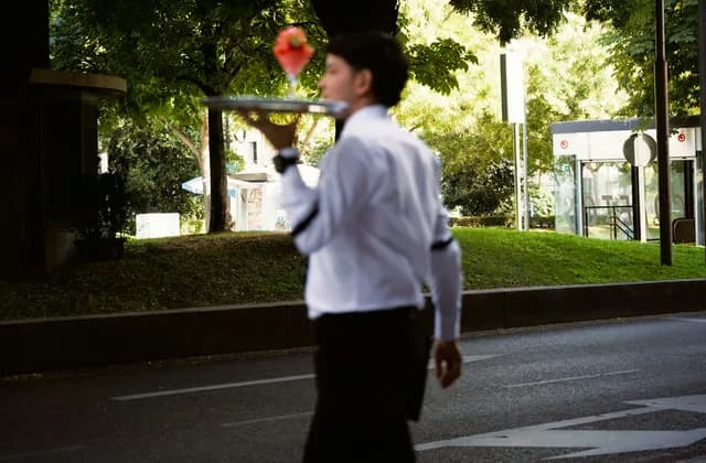 Beyaz gömlekli bir garson, tepside içecekle ağaçlı bir yolun karşısına geçiyor