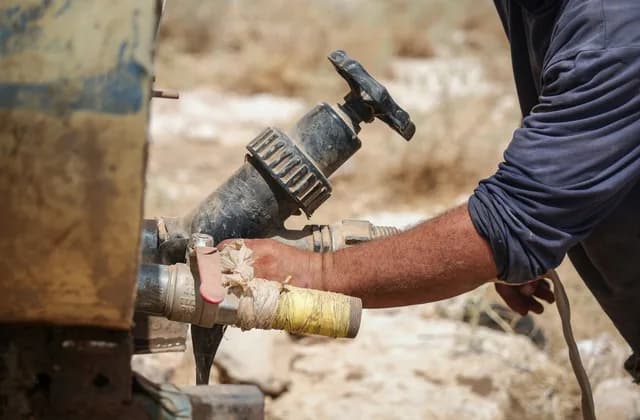 Kurak bir arazide su tankerinin vanasına bantla sarılmış hortum bağlayan bir el