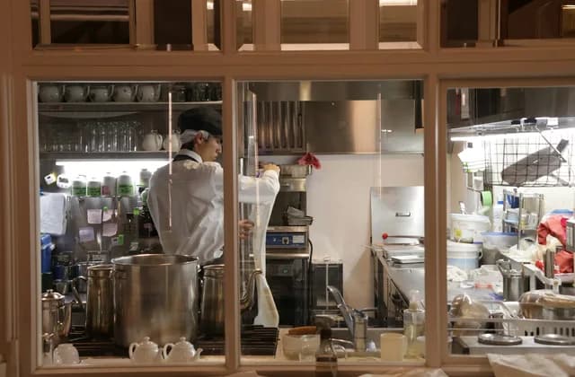 Ahşap çerçeveli camın ardında, büyük tencerelerin yanında beyaz ceketli bir aşçının çalıştığı restoran mutfağı