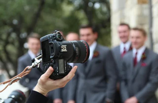 Mão segurando uma câmera fotográfica com padrinhos de casamento desfocados ao fundo