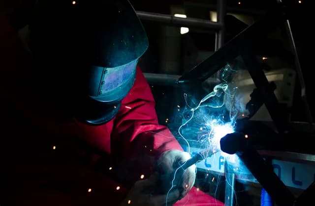 Kaynak maskesi ve eldivenli bir kaynakçı, atölyede metal bir konstrüksiyonu kaynatıyor; ark mavi ışık saçıyor