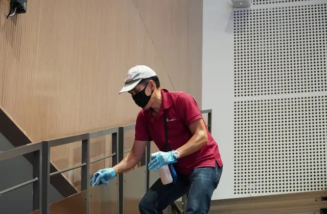 Maskeli ve eldivenli bir görevli, merdiven korkuluğunu sprey ve bezle temizliyor