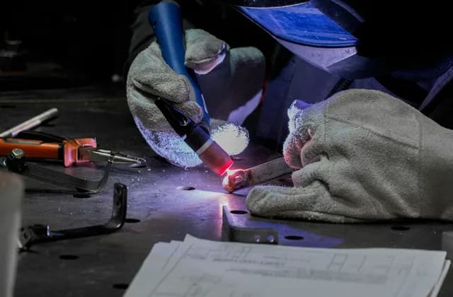 Deri eldivenli bir kaynakçı TIG torcu ve dolgu teliyle metal parçayı argon kaynağıyla birleştiriyor; tezgâhta teknik resim