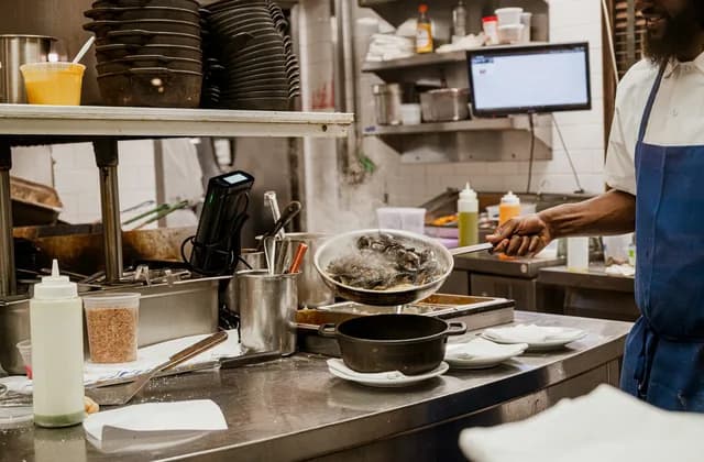 Posten einer Restaurantküche: Pfanne und Topf auf der Edelstahlablage, Teller und Quetschflaschen bereitgestellt, rechts ein Koch in weißer Jacke und blauer Schürze, im Hintergrund ein Monitor