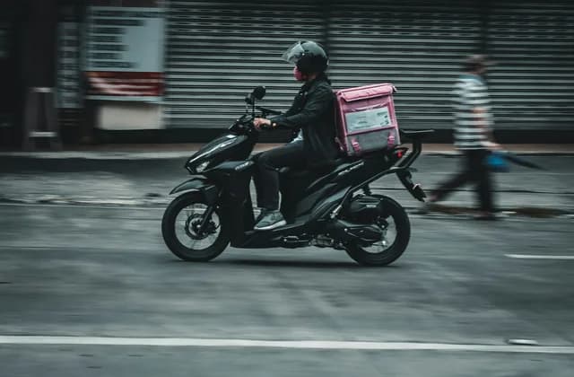 Kurierfahrer mit Helm auf einem schwarzen Motorroller mit hellem Transportkoffer am Heck, in Fahrt auf einer Straße; am Gehsteig dahinter geschlossene Rollläden